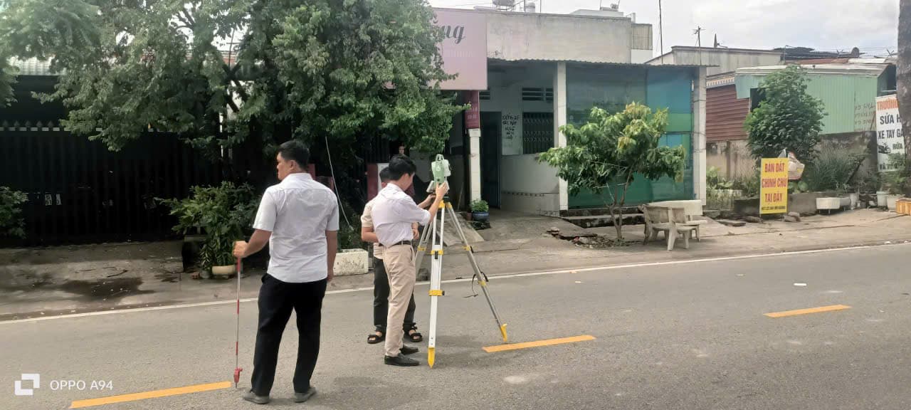 Hai nhân sự sử dụng máy toàn đạc và sào đo bên đường, phía trước các công trình nhà ở.